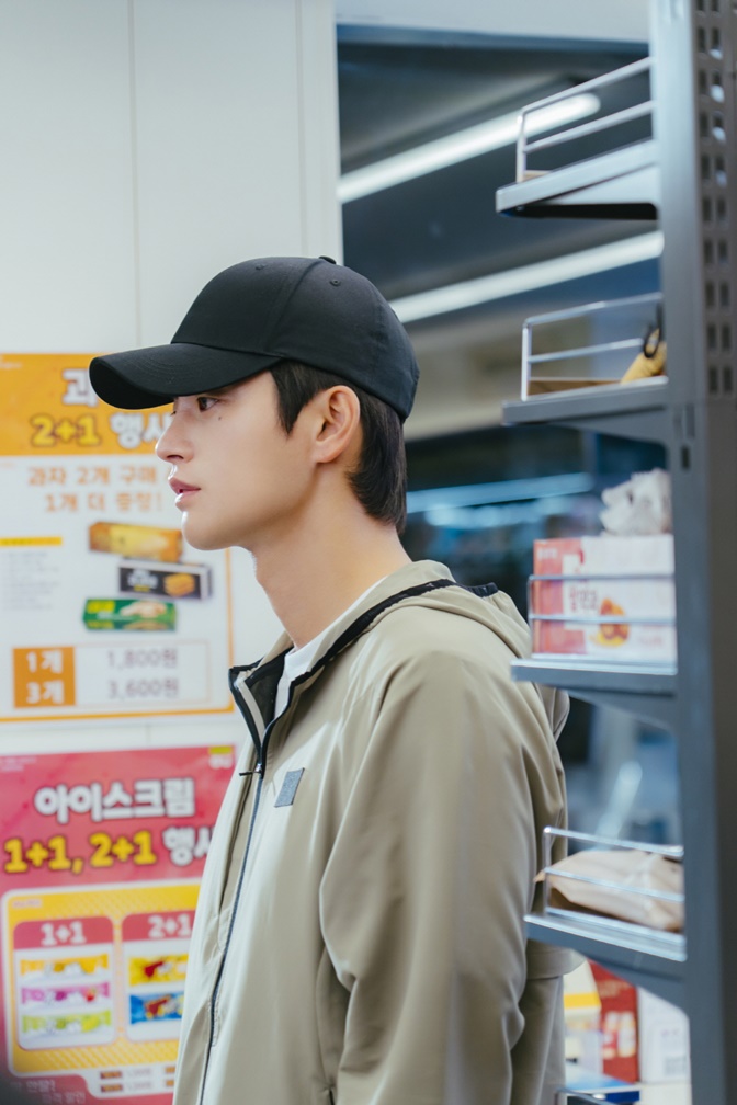 Seo In-guk como Kang Si-woo de boné e moletom observa produtos em loja de conveniência em Te vejo no trabalho!
