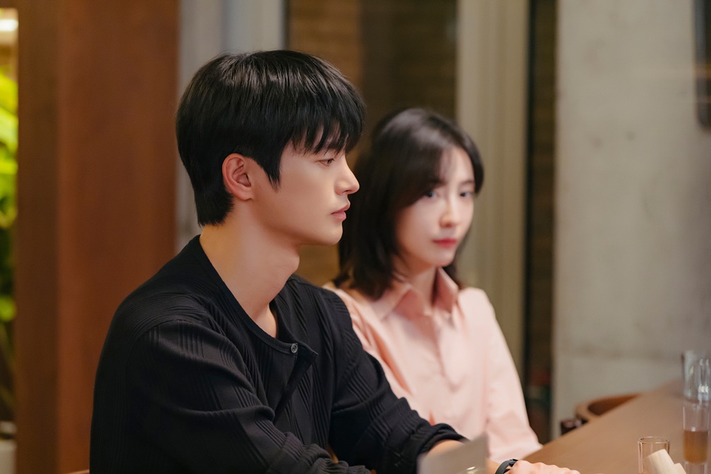 Seo In-guk como Kang Si-woo de suéter preto sentado ao lado de Park Ji-hyun como Cha Ji-yoon em bar em Te vejo no trabalho