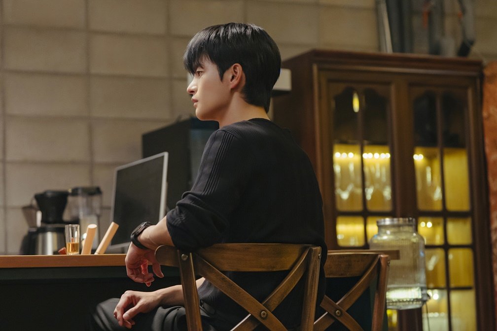 Seo In-guk como Kang Si-woo sentado relaxado em bar olhando para o lado em Te vejo no trabalho