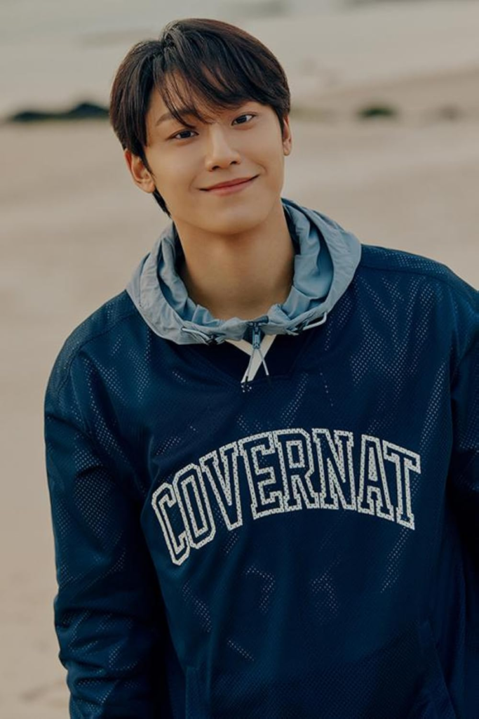 Lee Do-hyun sorri em foto promocional casual na praia vestindo moletom azul