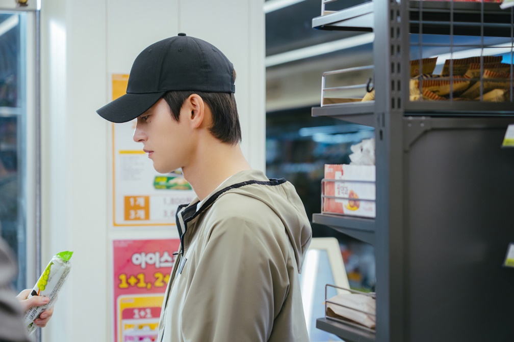Seo In-guk como Kang Si-woo de boné observa produto na mão de Park Ji-hyun em loja de conveniência em Te vejo no trabalho!