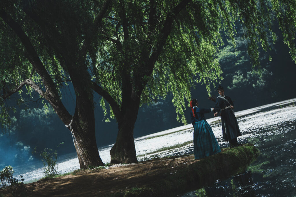 Nam Joo-hyuk e Roh Yoon-seo lado a lado junto a um lago se preparando para entrar no mundo dos fantasmas em A Leste do Palácio
