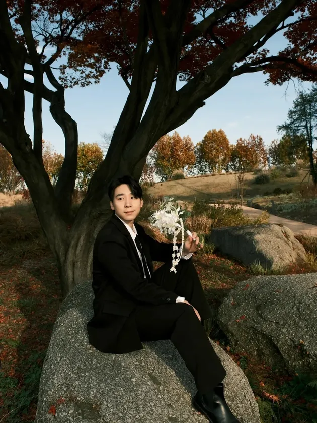O ator musical Lee Jun-woo sentado em uma grande rocha no outono, vestindo terno preto e segurando um buquê de flores brancas