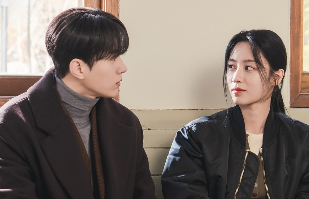 Kim Myung-soo e Kang Min-ah sentados lado a lado em um banco de madeira, se olhando, em still de Amor em Sincronia